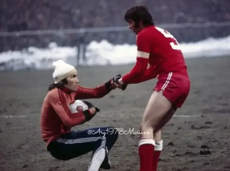 El Loco Hugo Orlando Gatti, con pantalones largos y gorrito por el frío extremo de Europa, es ayudado a levantarse por un futbolista polaco.